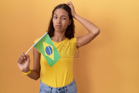 Téléchargez les photos : Jeune femme hispanique tenant drapeau brésilien confondre et se poser des questions. incertain avec doute, pensant la main sur la tête. concept coûteux. - en image libre de droit