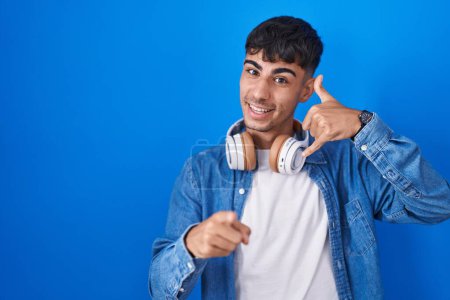 Photo for Young hispanic man standing over blue background smiling doing talking on the telephone gesture and pointing to you. call me. - Royalty Free Image