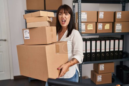 Photo for Young brunette woman working at small business ecommerce holding packages angry and mad screaming frustrated and furious, shouting with anger. rage and aggressive concept. - Royalty Free Image
