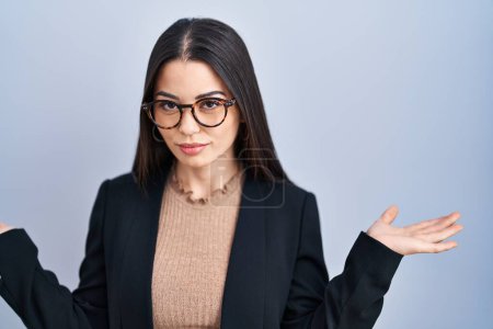 Photo for Young brunette woman standing over blue background clueless and confused expression with arms and hands raised. doubt concept. - Royalty Free Image