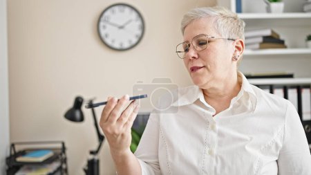 Téléchargez les photos : Femme d'affaires aux cheveux gris du moyen âge envoyant un message vocal par téléphone intelligent travaillant au bureau - en image libre de droit