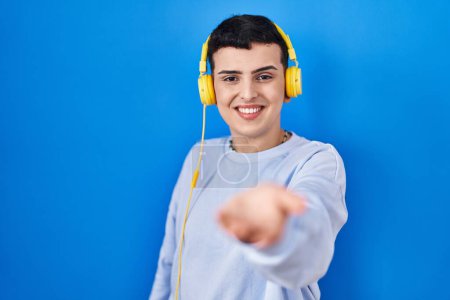 Téléchargez les photos : Personne non binaire écoutant de la musique à l'aide d'écouteurs souriant joyeuse offrant la main de la paume donnant assistance et acceptation. - en image libre de droit