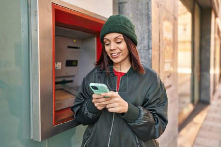 Téléchargez les photos : Jeune belle femme hispanique à l'aide d'un smartphone debout près du guichet de la banque à la rue - en image libre de droit