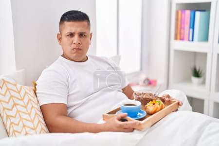Foto de Joven hombre hispano desayunando en la cama pensando actitud y expresión sobria buscando confianza en sí mismo - Imagen libre de derechos
