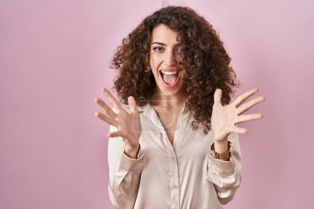 Téléchargez les photos : Hispanic woman with curly hair standing over pink background celebrating crazy and amazed for success with arms raised and open eyes screaming excited. winner concept - en image libre de droit