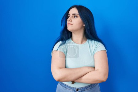 Téléchargez les photos : Jeune fille moderne avec les cheveux bleus debout sur fond bleu regardant sur le côté avec les bras croisés convaincu et confiant - en image libre de droit
