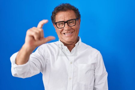 Photo for Middle age hispanic man standing over blue background smiling and confident gesturing with hand doing small size sign with fingers looking and the camera. measure concept. - Royalty Free Image