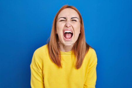 Foto de Young woman standing over blue background angry and mad screaming frustrated and furious, shouting with anger. rage and aggressive concept. - Imagen libre de derechos