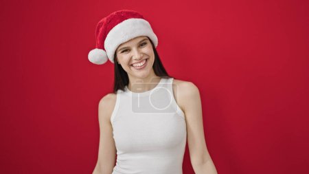 Foto de Joven hermosa mujer hispana sonriendo confiada usando sombrero de Navidad sobre fondo rojo aislado - Imagen libre de derechos