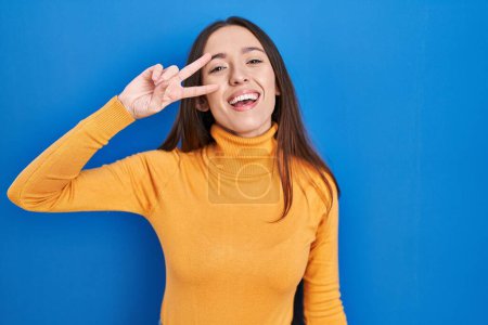 Foto de Young brunette woman standing over blue background doing peace symbol with fingers over face, smiling cheerful showing victory - Imagen libre de derechos