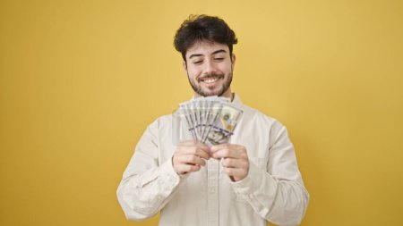Téléchargez les photos : Jeune homme hispanique souriant confiant comptant les dollars sur fond jaune isolé - en image libre de droit