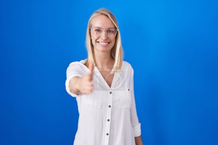 Téléchargez les photos : Young caucasian woman standing over blue background smiling friendly offering handshake as greeting and welcoming. successful business. - en image libre de droit