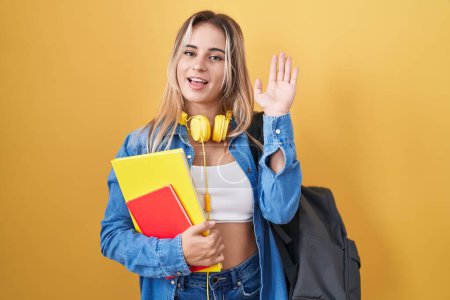 Téléchargez les photos : Jeune femme blonde portant un sac à dos étudiant et tenant des livres renonçant à dire bonjour heureux et souriant, geste de bienvenue amical - en image libre de droit