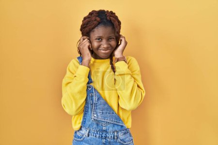Foto de Mujer africana de pie sobre fondo amarillo cubriendo las orejas con los dedos con expresión molesta por el ruido de la música fuerte. concepto de sordo. - Imagen libre de derechos