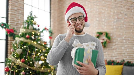 Téléchargez les photos : Homme hispanique portant un chapeau de Noël tenant un cadeau parlant au téléphone à la maison - en image libre de droit