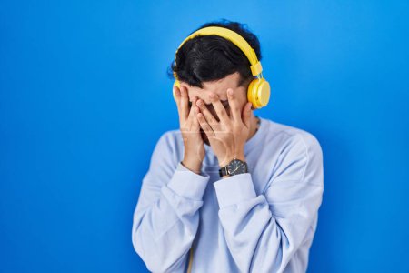 Foto de Persona no binaria escuchando música usando auriculares con expresión triste cubriendo la cara con las manos mientras llora. concepto de depresión. - Imagen libre de derechos