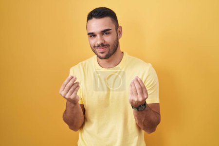Téléchargez les photos : Jeune homme hispanique debout sur fond jaune faisant geste d'argent avec les mains, demandant le paiement du salaire, entreprise millionnaire - en image libre de droit