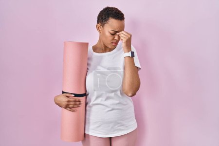 Photo for Beautiful african american woman holding yoga mat tired rubbing nose and eyes feeling fatigue and headache. stress and frustration concept. - Royalty Free Image