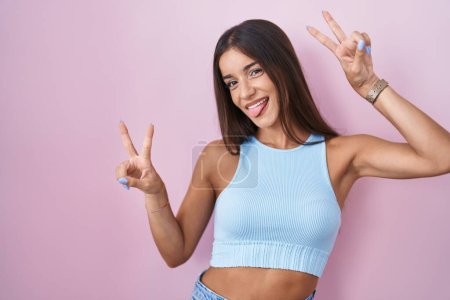 Photo for Young brunette woman standing over pink background smiling with tongue out showing fingers of both hands doing victory sign. number two. - Royalty Free Image