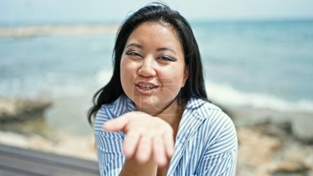 Téléchargez les photos : Jeune femme chinoise souffle un baiser avec la main assise sur un banc au bord de la mer - en image libre de droit