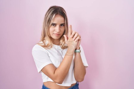 Photo for Young blonde woman standing over pink background holding symbolic gun with hand gesture, playing killing shooting weapons, angry face - Royalty Free Image
