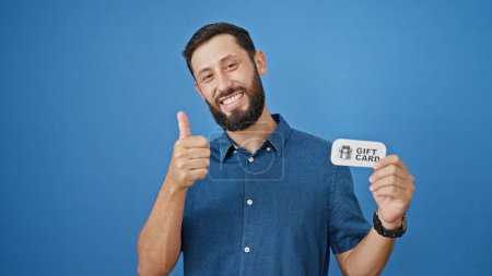 Foto de Joven hombre hispano sonriendo confiado sosteniendo tarjeta de regalo haciendo gesto de pulgar hacia arriba sobre fondo azul aislado - Imagen libre de derechos