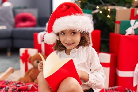 Téléchargez les photos : Adorable hispanique fille lecture livre assis sur le sol par arbre de Noël à la maison - en image libre de droit