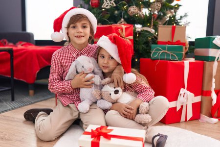 Foto de Hermano y hermana abrazándose el uno al otro sentados en el suelo por el árbol de Navidad en casa - Imagen libre de derechos