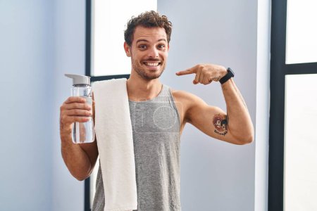 Foto de Joven hombre hispano usando ropa deportiva bebiendo agua buscando confiado con sonrisa en la cara, señalándose con los dedos orgullosos y felices. - Imagen libre de derechos