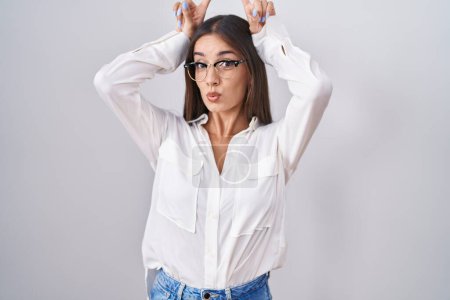 Téléchargez les photos : Jeune femme brune portant des lunettes faisant un geste drôle avec le doigt sur la tête comme des cornes de taureau - en image libre de droit