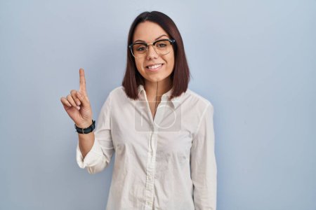 Foto de Joven mujer hispana de pie sobre fondo blanco mostrando y señalando con el dedo número uno mientras sonríe confiada y feliz. - Imagen libre de derechos