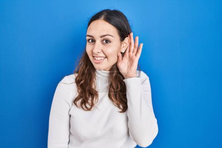 Téléchargez les photos : Young hispanic woman standing over blue background smiling with hand over ear listening an hearing to rumor or gossip. deafness concept. - en image libre de droit