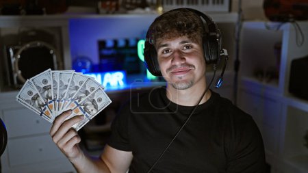 Photo for Handsome young hispanic man streaming late-night gaming session while flashing dollars, savoring his virtual game success from his home office. - Royalty Free Image