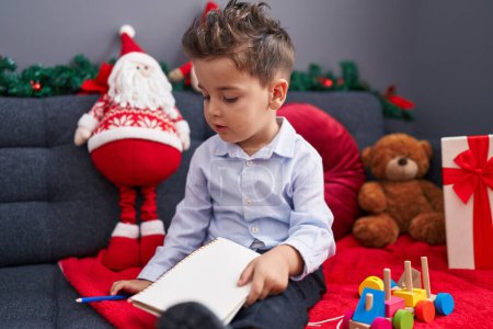 Photo for Adorable hispanic toddler writing letter to santa claus sitting on sofa by christmas decoration at home - Royalty Free Image