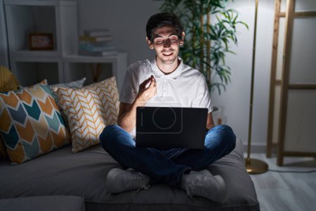 Téléchargez les photos : Jeune homme hispanique utilisant un ordinateur portable à la maison la nuit faire un geste d'argent avec les mains, demander le paiement du salaire, entreprise millionnaire - en image libre de droit