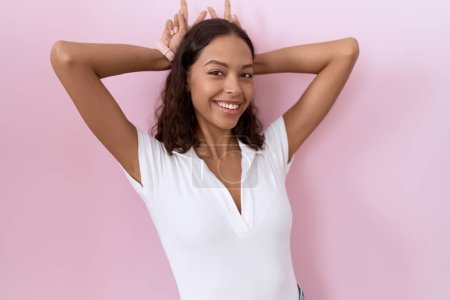 Foto de Mujer hispana joven vistiendo camiseta blanca casual posando divertida y loca con los dedos en la cabeza como orejas de conejo, sonriendo alegre - Imagen libre de derechos
