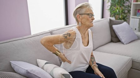 Photo for Unhappy grey-haired senior woman suffers severe backache, sitting on home sofa. worried, mature female adults grapple with serious spine problem indoors in middle age. - Royalty Free Image