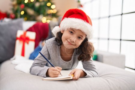 Foto de Adorable chica china escribiendo en el cuaderno acostado en el sofá por el árbol de Navidad en casa - Imagen libre de derechos