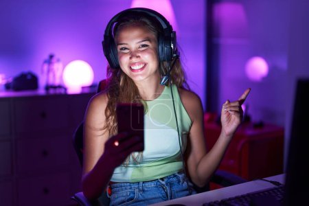 Téléchargez les photos : Jeune femme hispanique jouant à des jeux vidéo avec smartphone souriant heureux pointant avec la main et le doigt sur le côté - en image libre de droit