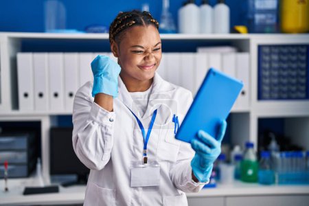 Téléchargez les photos : Femme afro-américaine avec des tresses travaillant au laboratoire scientifique faire appel vidéo avec tablette criant fier, célébrant la victoire et le succès très excité avec le bras levé - en image libre de droit