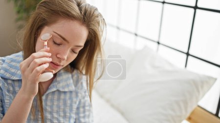 Foto de Mujer rubia joven masajeando la cara con la piel gimnasio sentado en la cama en el dormitorio - Imagen libre de derechos