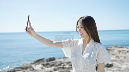 Foto de Joven mujer china sonriendo confiada haciendo selfie por el teléfono inteligente en la playa - Imagen libre de derechos
