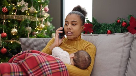 Foto de Madre e hijo celebrando la Navidad hablando en el teléfono inteligente mientras amamanta al bebé en casa - Imagen libre de derechos