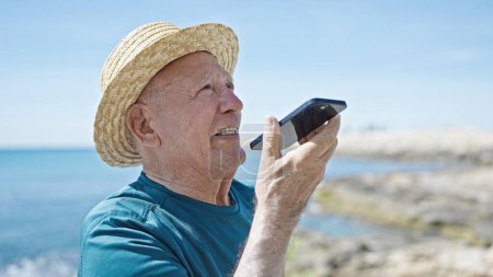 Téléchargez les photos : Homme âgé aux cheveux gris touriste portant un chapeau d'été envoyant un message vocal par smartphone au bord de la mer - en image libre de droit