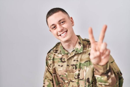 Foto de Joven vestido con camuflaje uniforme del ejército sonriendo mirando a la cámara mostrando los dedos haciendo señal de victoria. número dos. - Imagen libre de derechos
