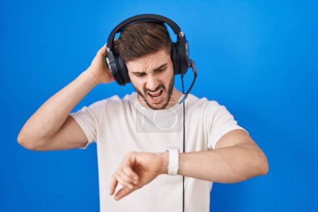 Téléchargez les photos : Homme hispanique avec barbe écoutant de la musique portant des écouteurs regardant l'heure de la montre inquiet, peur de se retrouver en retard - en image libre de droit