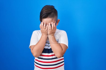 Foto de Joven niño hispano de pie sobre fondo azul con expresión triste cubriendo la cara con las manos mientras llora. concepto de depresión. - Imagen libre de derechos