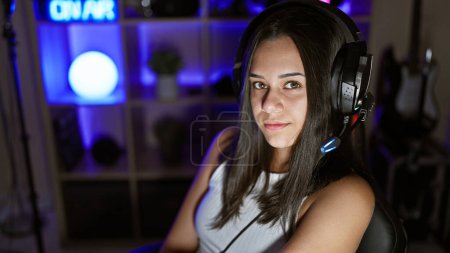 Photo for Young beautiful hispanic woman streamer sitting on table with serious face at gaming room - Royalty Free Image