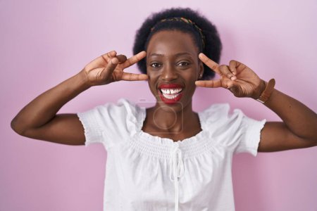 Téléchargez les photos : African woman with curly hair standing over pink background doing peace symbol with fingers over face, smiling cheerful showing victory - en image libre de droit