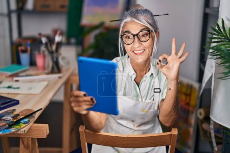 Foto de Mujer de mediana edad con el pelo gris sentado en el estudio de arte utilizando la tableta haciendo signo bien con los dedos, sonriendo gesto amistoso excelente símbolo - Imagen libre de derechos